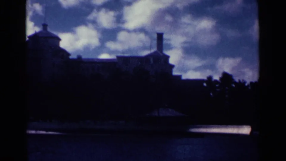 1963 Nassau home-movie frame showing a shadowed waterfront structure, palm trees, water, and a clouded blue-purple sky.