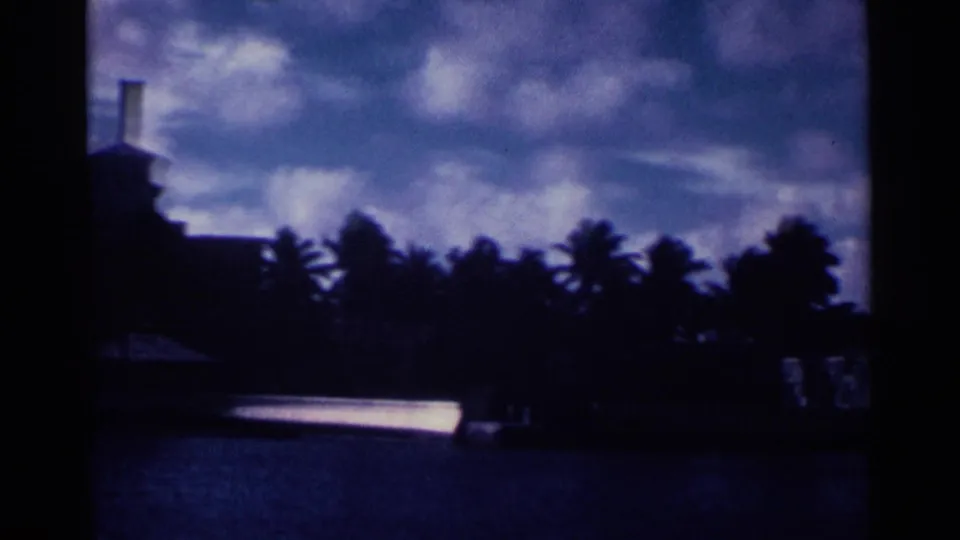 1963 Nassau waterfront view with a dark building edge, palm silhouettes, water, and mottled clouds in blue evening-toned film color.