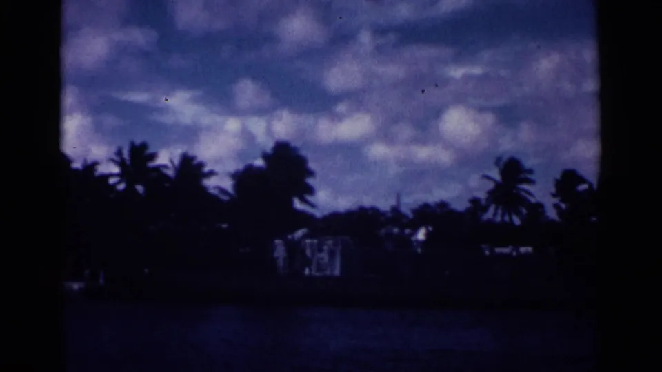 1963 Nassau home-movie frame showing a low palm-lined shore, small light buildings, dark water, and a textured cloudy sky.