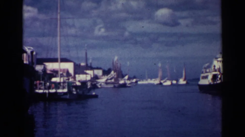 1963 Nassau harbor view with small sailboats, waterfront buildings, calm blue water, and a larger work boat partly entering frame.