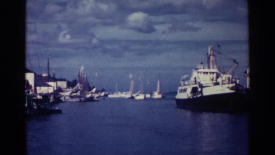 1963 Nassau harbor frame showing a white and dark work boat near the right side, with sailboats and waterfront buildings in the distance.
