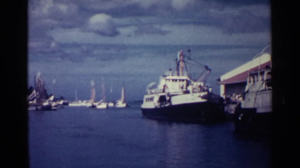 1963 Nassau harbor scene with sailboats across the water, a larger fishing or work boat, and a docked structure along the right edge.