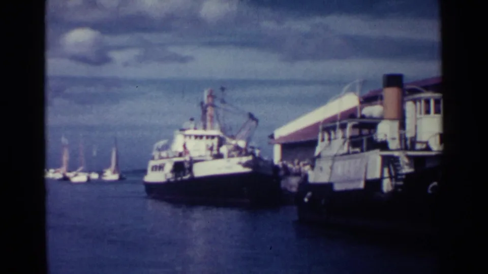 1963 Nassau harbor view with a large white and dark vessel near a dock, sailboats in the distance, and cloudy sky above calm water.