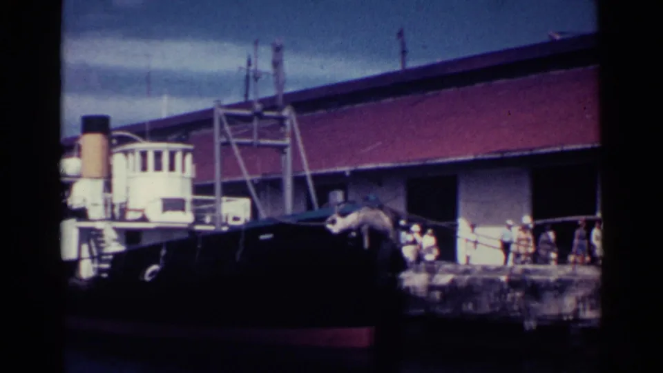 Workers gather on a Nassau pier beside a moored dark-hulled boat and a red-roofed working building in 1963 footage.