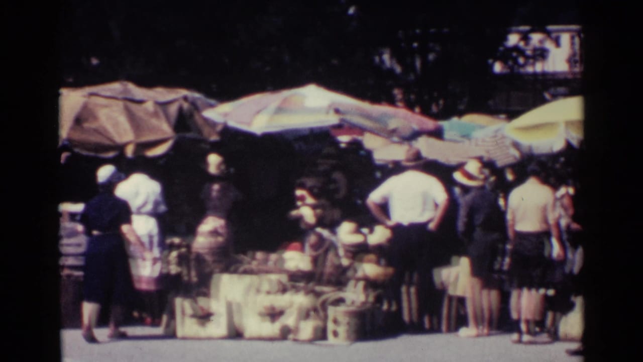 Shoppers stand near umbrella-covered market stalls with goods arranged along the roadside.