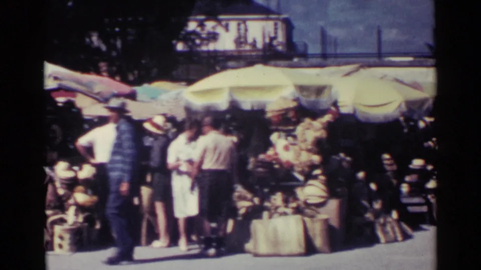 Nassau market visitors move past umbrella-covered stalls, with hats and light clothing visible in the crowd.