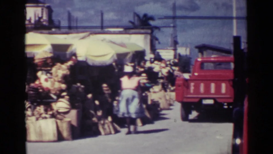 A red pickup appears beside Nassau market stalls while shoppers move near umbrellas and stacked goods.