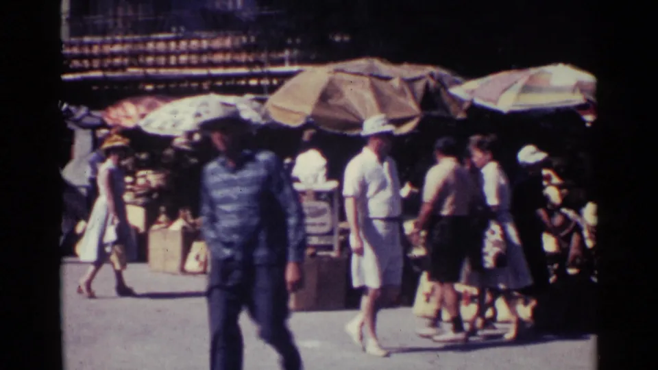 People in 1960s clothing walk past umbrella-covered Nassau market stalls with goods stacked near the street.