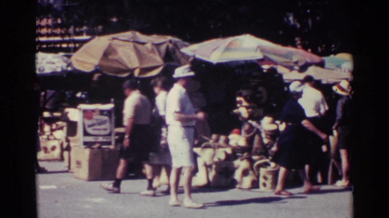 Shoppers browse an outdoor market with umbrellas, baskets, and vendor displays.