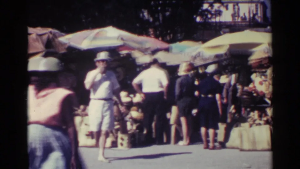 Outdoor Nassau market view with a man in light clothing, nearby shoppers, baskets, and shaded vendor tables.