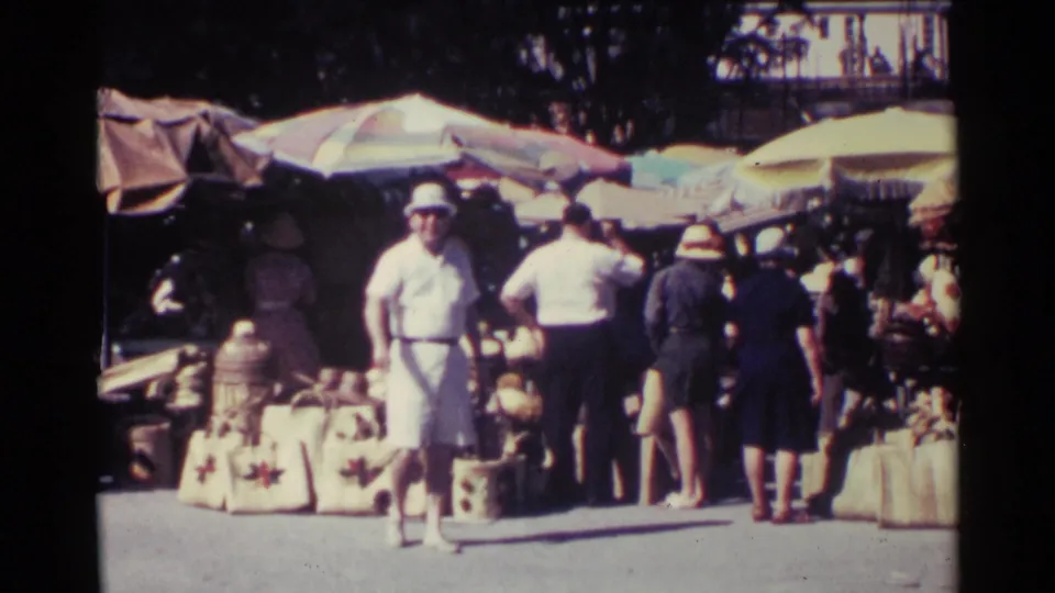 1963 Nassau outdoor market with shoppers seen from behind and stalls displaying baskets beneath large sun umbrellas.