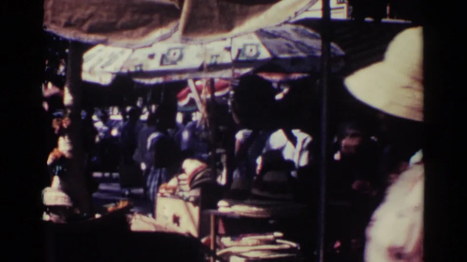 Dimmer market view in Nassau showing shoppers under a canopy with baskets and vendor goods in partial shade.