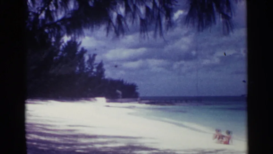 1963 Nassau beach frame with white sand, turquoise water, palm-shadowed foreground, and small seated beachgoers near the waterline.