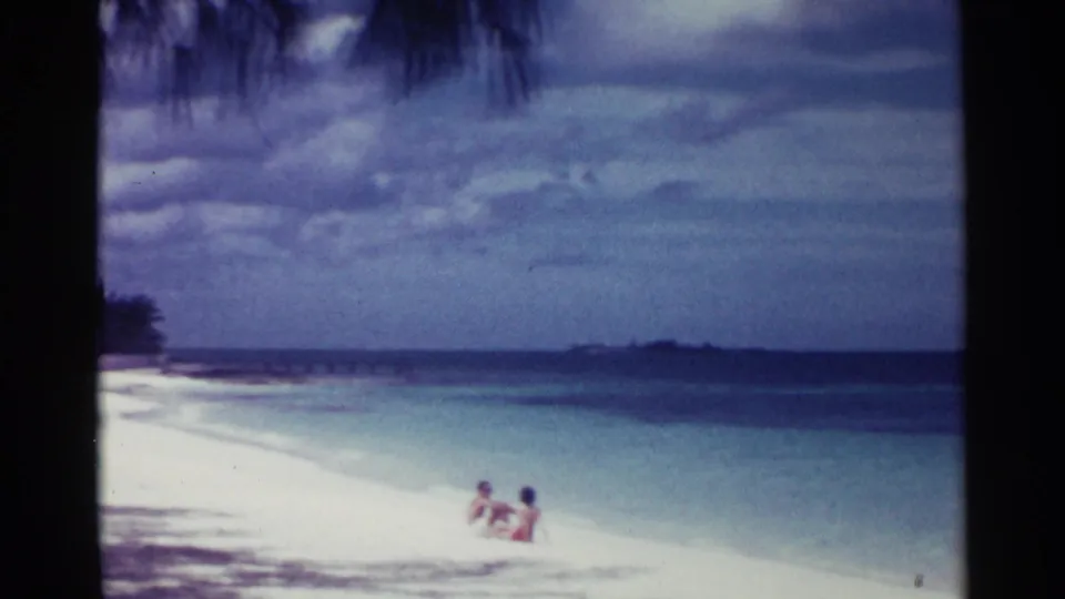Three people sit together on a sunlit Nassau beach in 1963, with pale sand, turquoise water, and a low island horizon behind them.