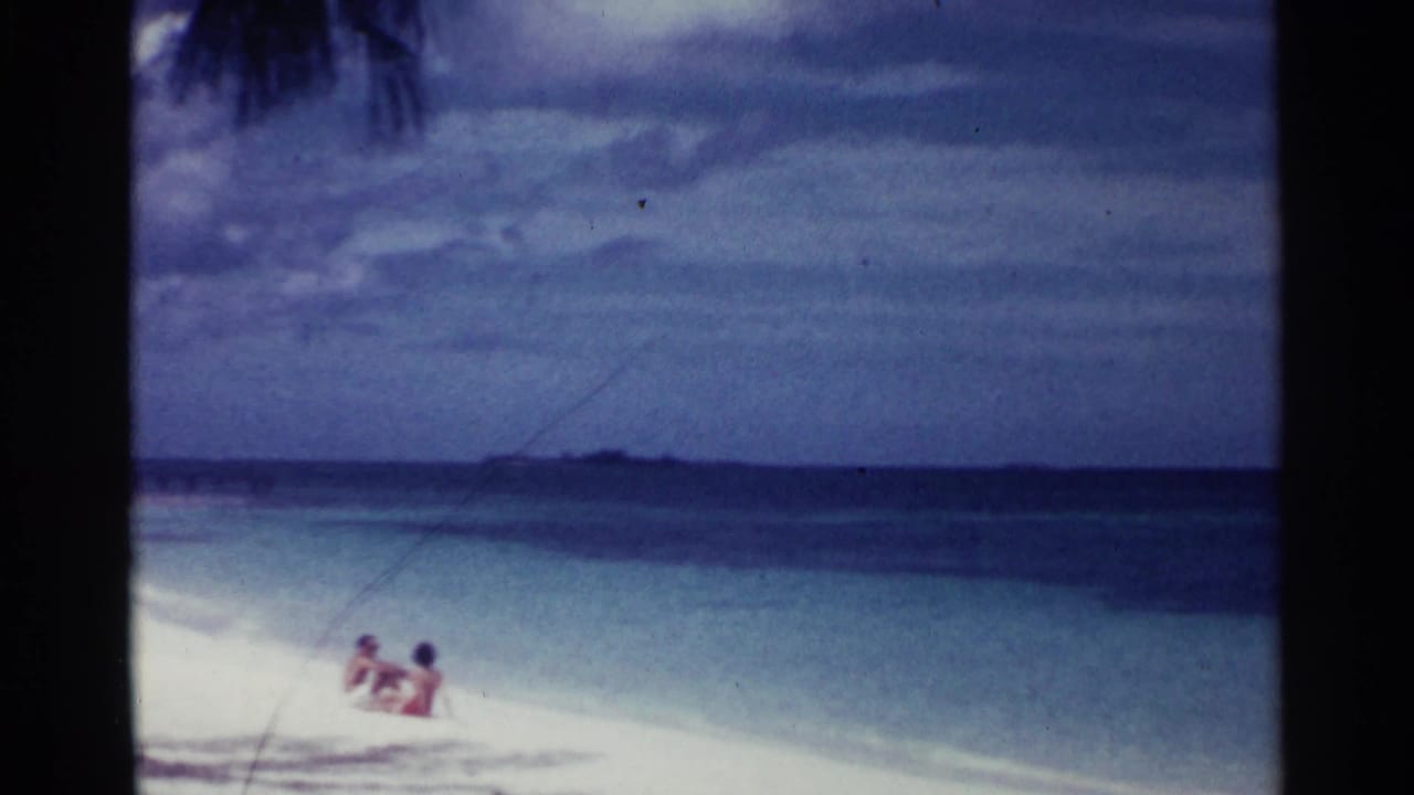 Two people sit or recline near the surf on a pale beach beside blue water.