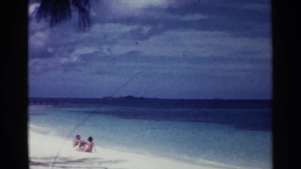 1963 Nassau beach home-movie frame with two distant people relaxing at the waterline under a cloudy sky.