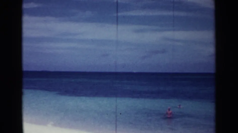 1963 Nassau beach frame with distant swimmers wading in shallow turquoise water below a cloudy sky.