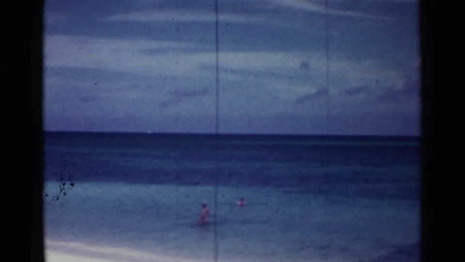 1963 Nassau beach home-movie frame showing distant swimmers in calm water with visible film wear and borders.