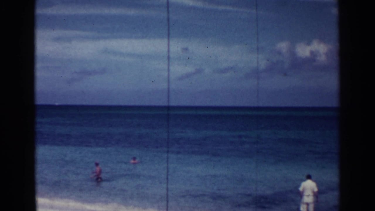 Man near the shoreline with swimmers in shallow ocean water under a cloudy sky.