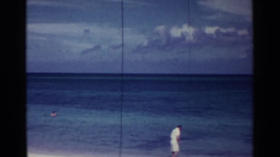 A distant man in light clothing walks beside the surf in Nassau, Bahamas, with swimmers in the ocean beyond.