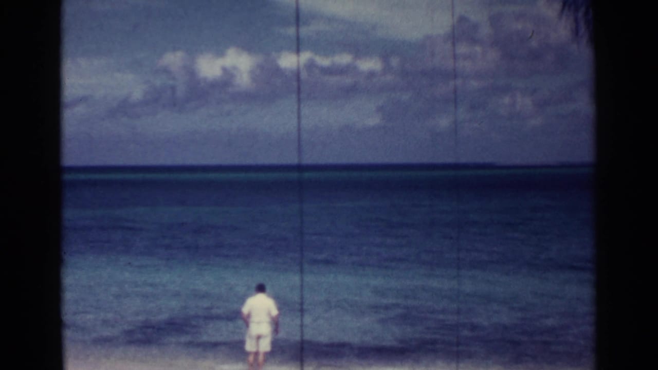 Man in white clothing stands near the water facing a blue sea under a cloudy sky.