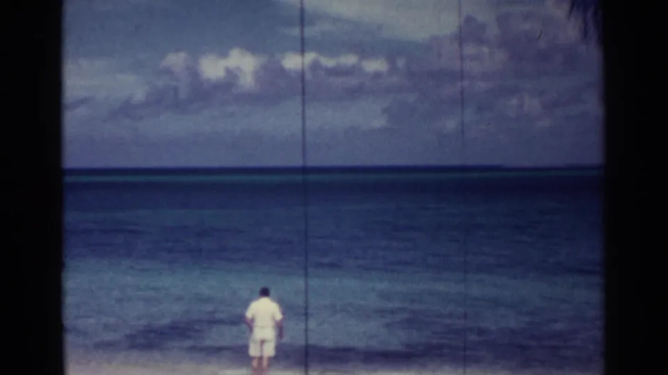 1963 Nassau home-movie frame showing a man in white standing on a beach and looking across blue water beneath clouds.