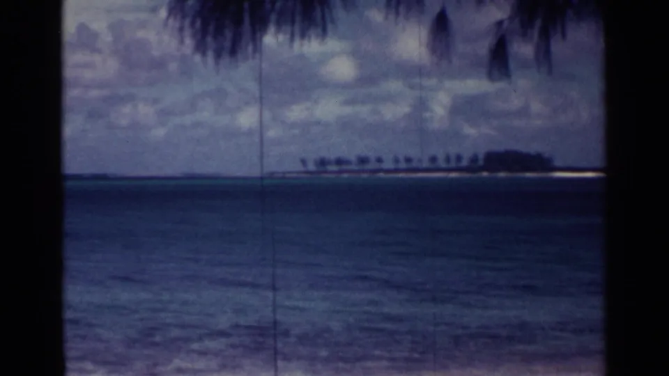 1963 Nassau 8mm frame with blue sea, a low palm-lined island, cloudy sky, and dark palm fronds overhead.