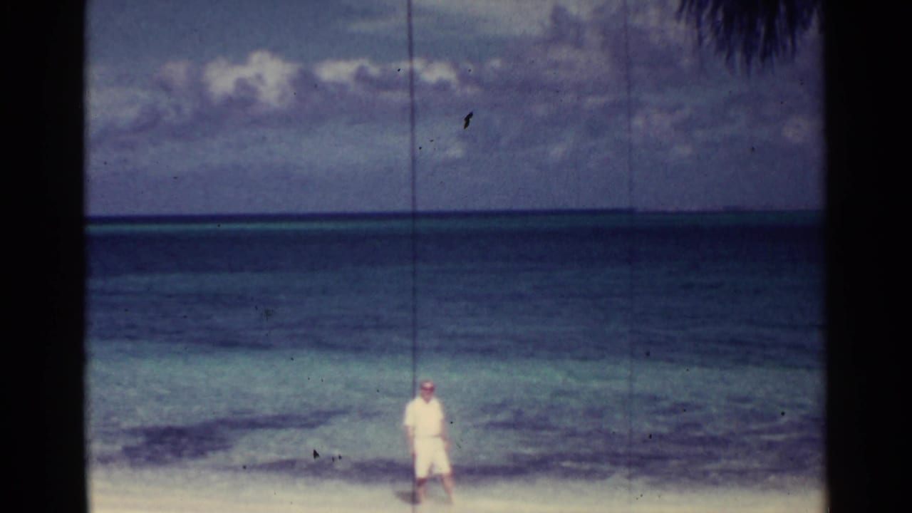 Man in white clothing standing near the waterline beneath a cloudy tropical sky.