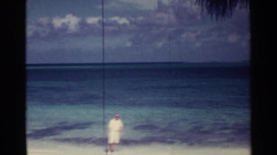 A man in white clothing stands on a Nassau beach near calm blue-green water in a soft 1963 8mm frame.