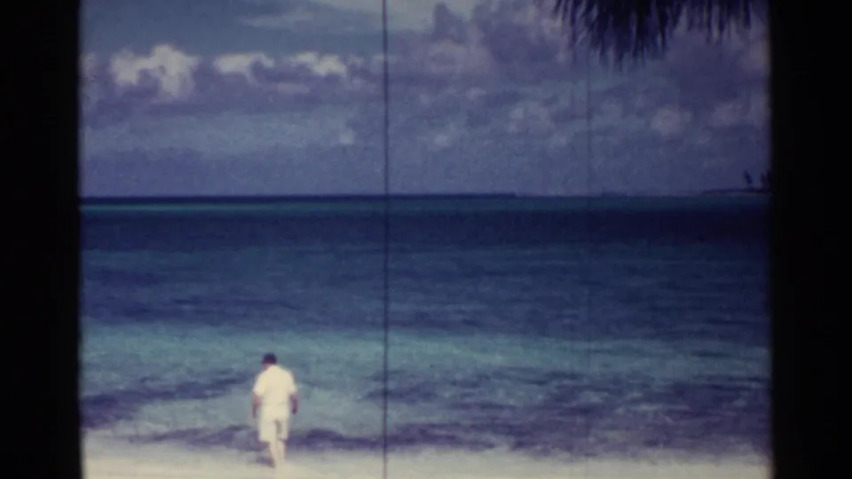 The lone beachgoer appears turned partly away on the Nassau shoreline, framed by ocean bands and cloudy sky in 1963 8mm footage.