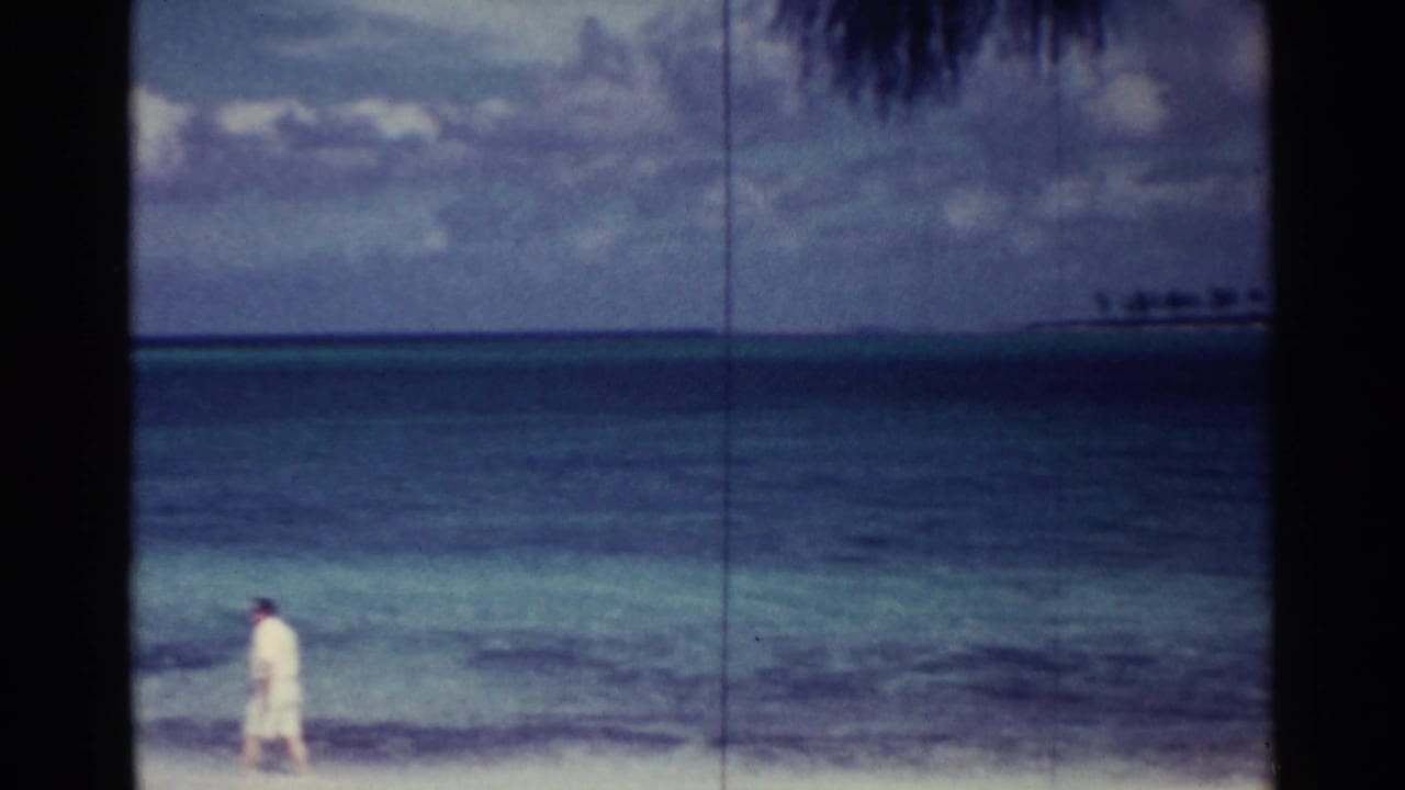 Lone person in light clothing walking near the edge of a tropical beach with ocean and distant land behind.