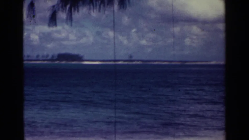 Nassau 1963 home-movie frame looking across dark blue water toward a low island or sandbar, with clouds and palm leaves at the top.