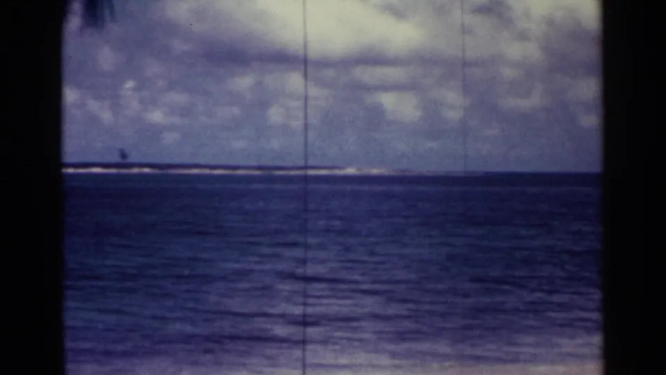 1963 Nassau beach footage frame showing open water, a low surf line, cloudy sky, and faint distant land through blue-purple 8mm color shift.