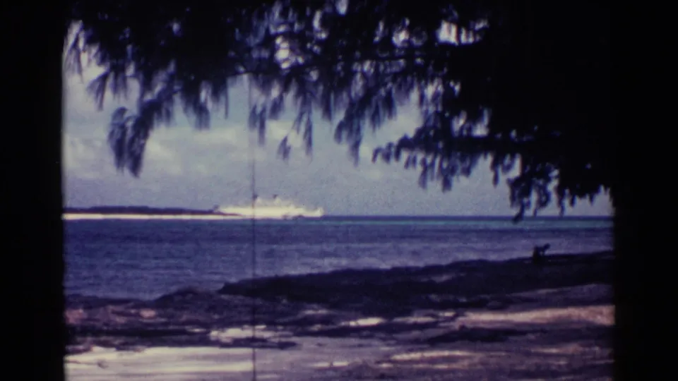 1963 Nassau coastal frame showing palm silhouettes, beach edge, open water, cloudy sky, and a distant white cruise liner near the horizon.