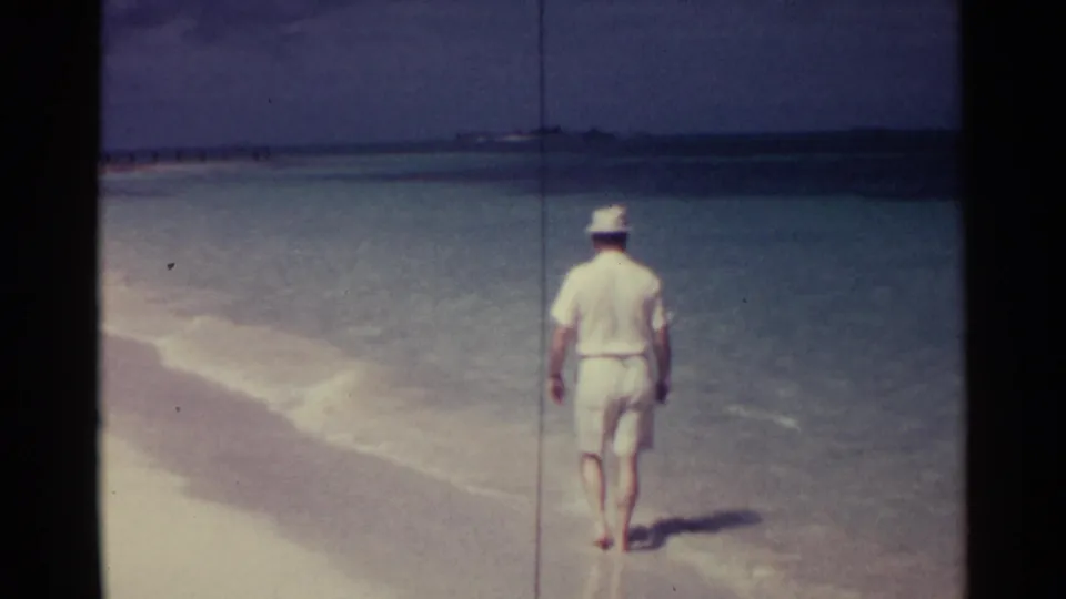 In Nassau, Bahamas, a man in white shirt, shorts, and hat walks barefoot away from camera along a quiet turquoise shoreline.