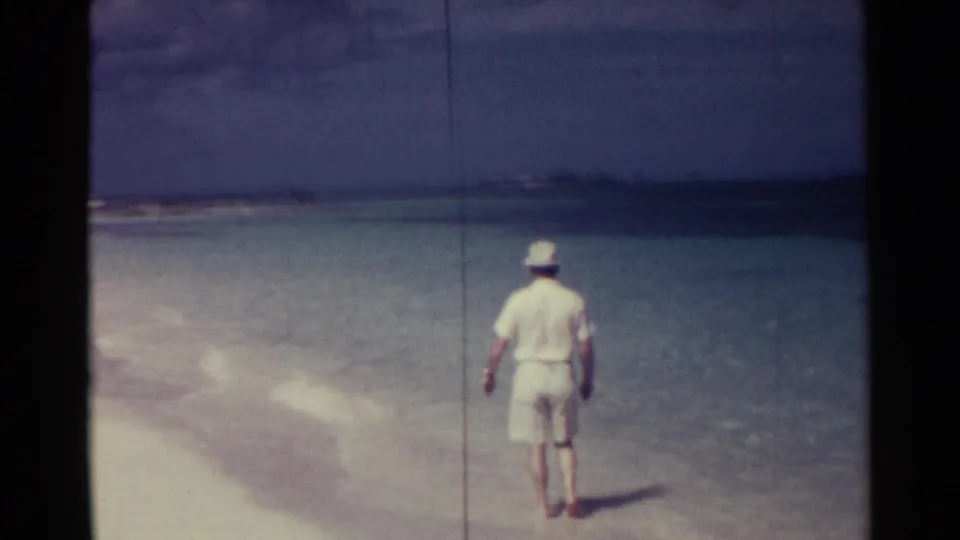 A 1963 Nassau vacation frame shows the man continuing barefoot along pale sand beside shallow blue-green water under a cloudy sky.