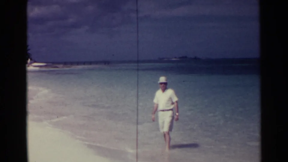 Near the Nassau shoreline, the man in white clothing and hat faces the camera briefly, with turquoise water and distant land behind him.