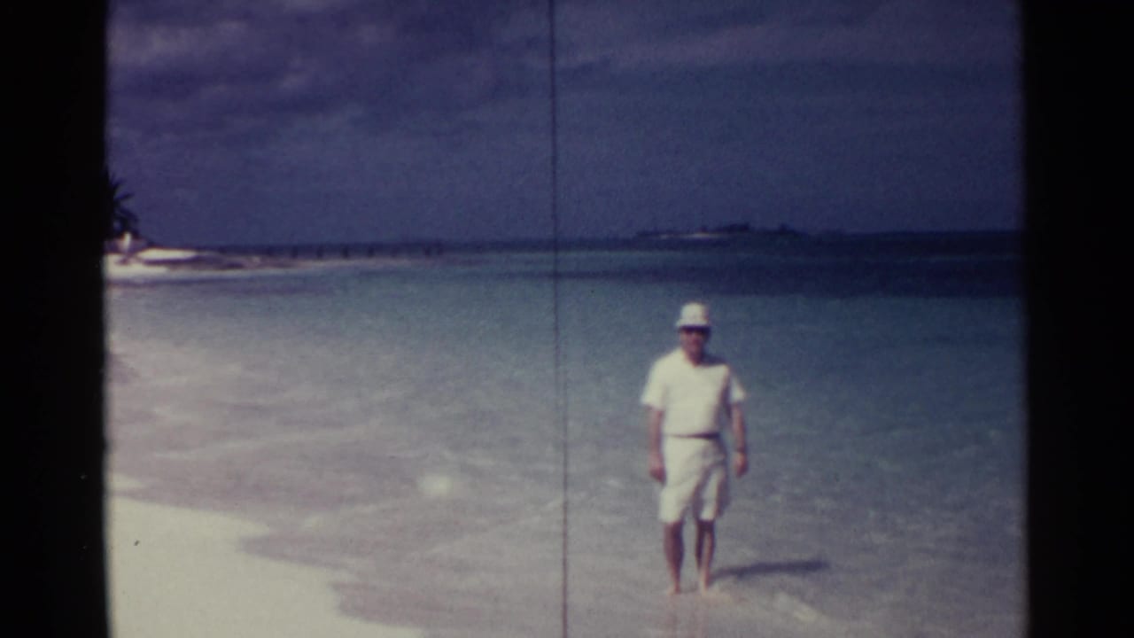 Man in white clothing and hat walking through shallow turquoise water near a sandy beach.