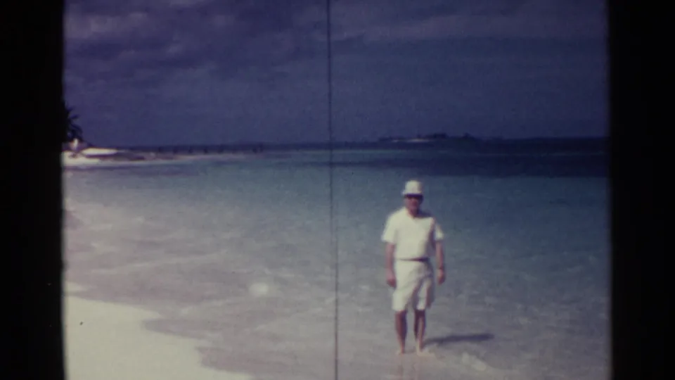 Adult man in white summer clothes and hat walks through shallow water at a Nassau beach, consistent with 1963 home-movie footage.