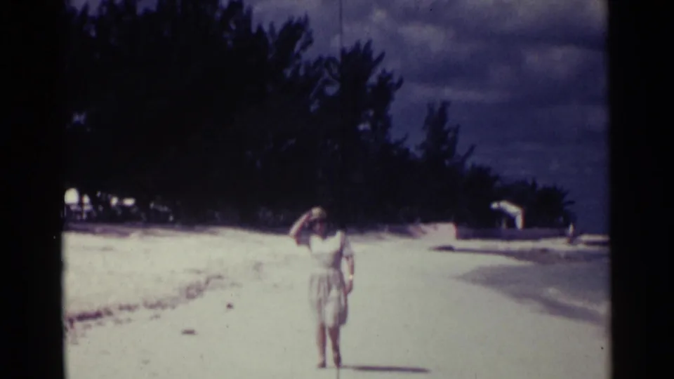 Adult woman in a light dress and hat walks toward the camera along a Nassau beach, with trees and shoreline behind her.