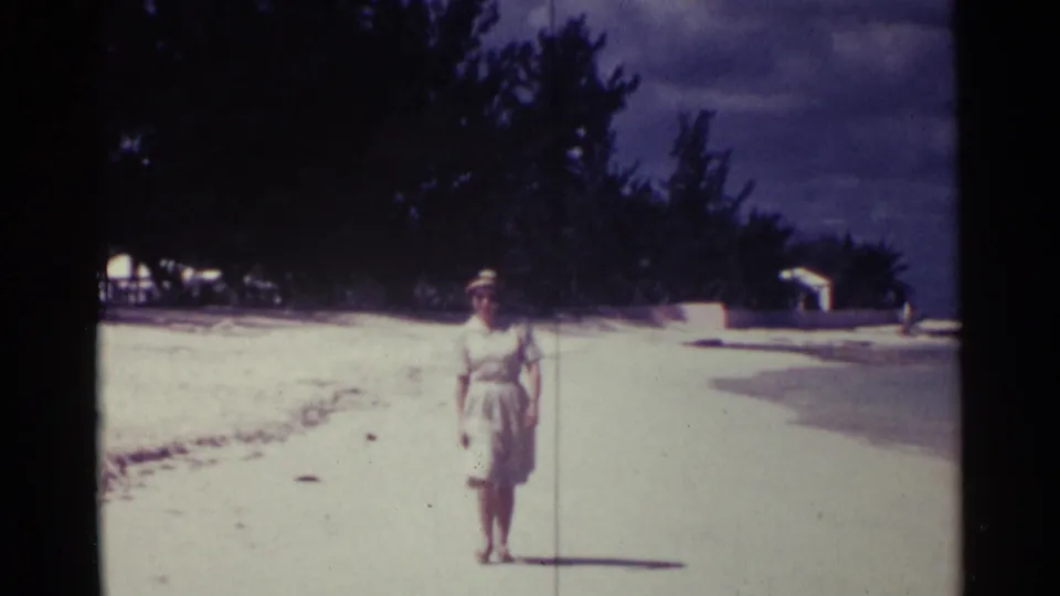 Adult woman in light summer clothing approaches the camera on a Nassau shoreline in faded 8mm color.