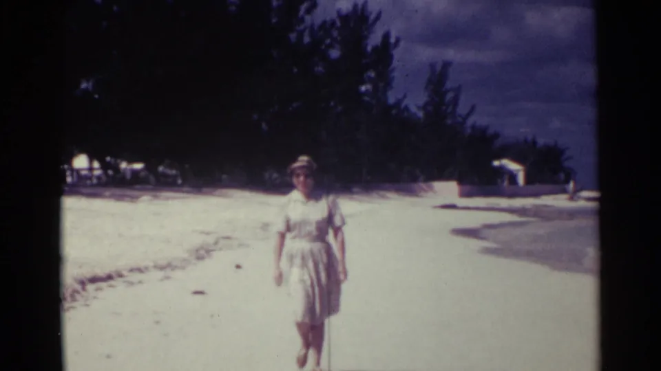 Adult woman in a hat and dress walks on the pale Nassau beach, with shoreline water, trees, and small structures behind her.