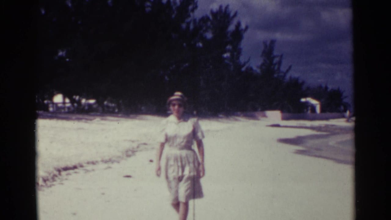 Woman in a light dress, hat, and sunglasses walking on a sandy beach.