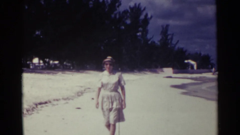 In Nassau, Bahamas, 1963, a woman in a pale dress, brimmed hat, and sunglasses walks toward the camera on a sandy beach.
