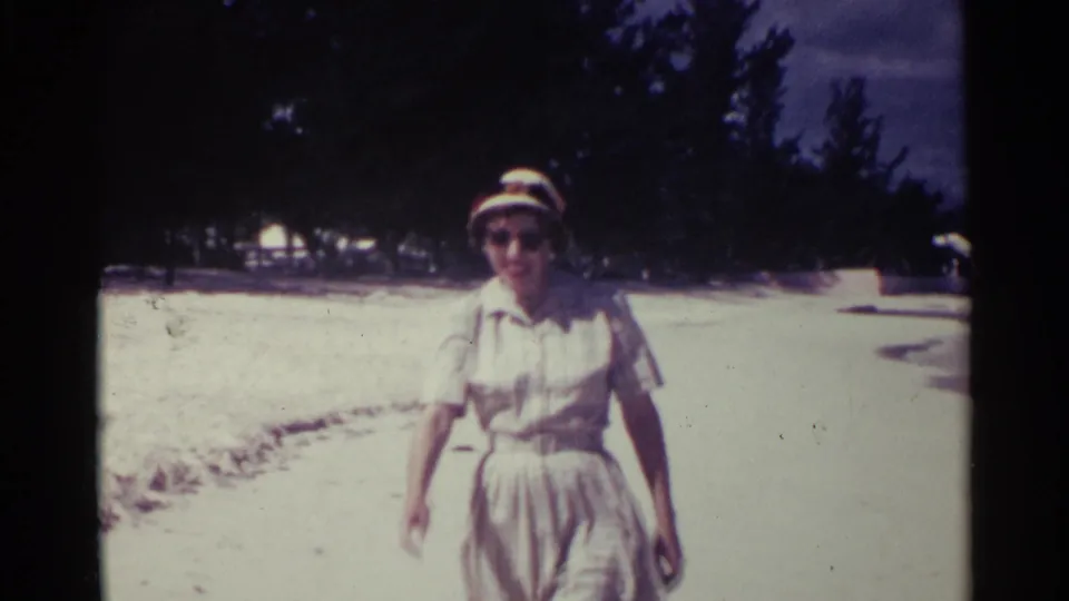 A closer 1963 beach view shows the woman in a belted pale dress, hat, and sunglasses walking toward the handheld camera.