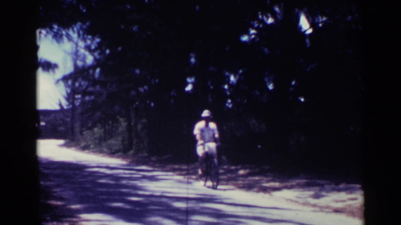 A man in a hat rides a bicycle toward the camera on a shaded road.
