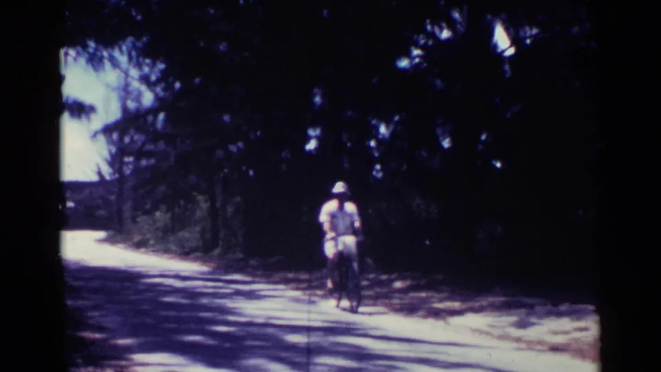 A man wearing a wide-brimmed hat rides a bicycle toward the camera on a tree-shaded Nassau road in 1963.