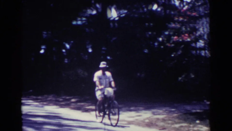 The rider comes closer on the rural Nassau road, with his hat, light clothing, bicycle frame, and surrounding trees visible.