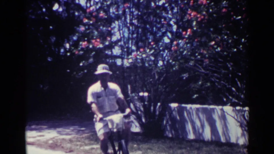 Near the end of the 1963 Nassau clip, the bicyclist passes close to the camera with a flowering bush and roadside wall or edge nearby.