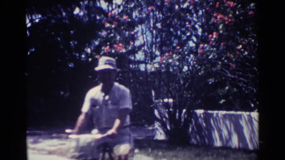 In 1963 Nassau, Bahamas, a woman in a white dress rides a bicycle near flowering trees and a low roadside wall.